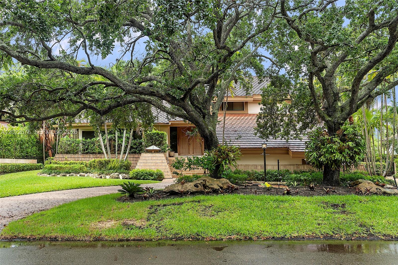 A two-story house with a brown tile roof is framed by large, mature oak trees — 7244 E Lago Dr, Coral Gables FL 33143, CORAL GABLES, FL