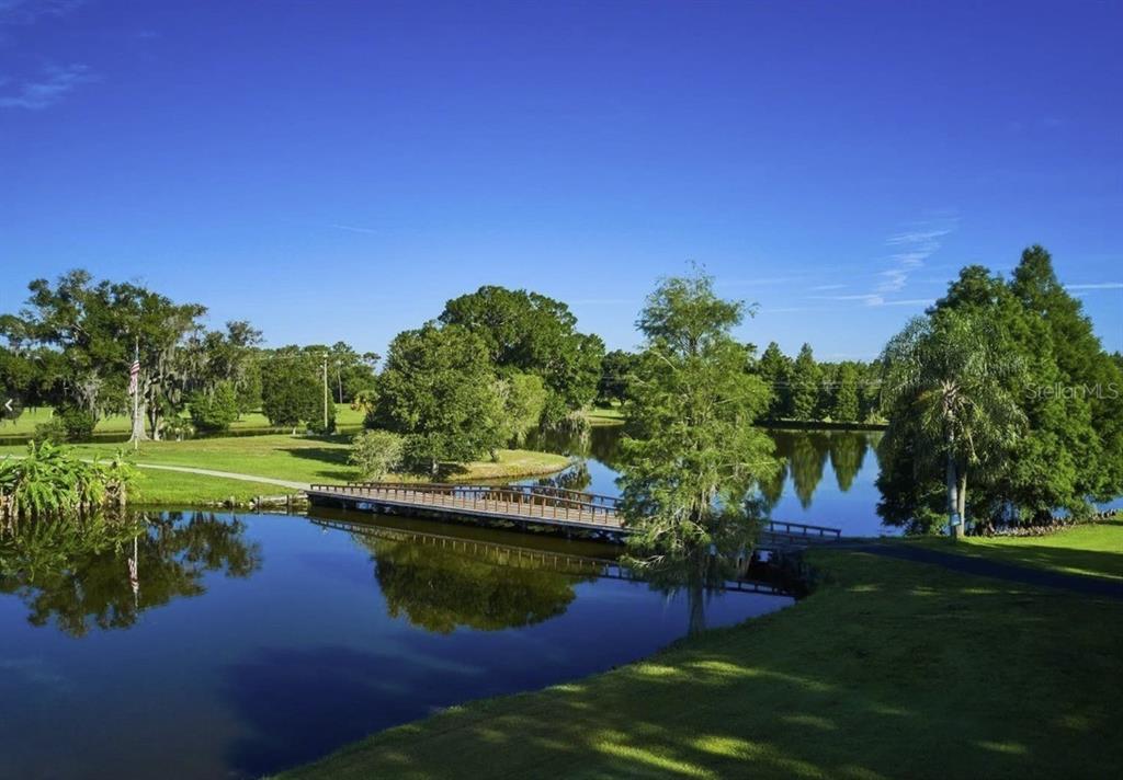 A wooden footbridge crosses a calm pond surrounded by green lawns and mature trees — 211 VALENCIA DRIVE, INDIAN LAKE ESTATES, FL