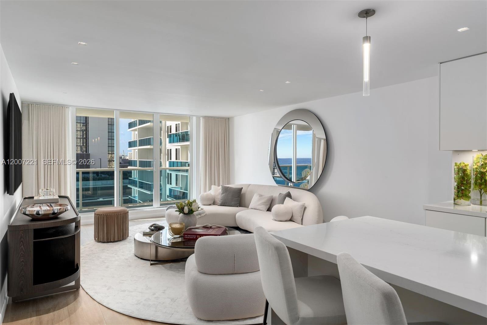 A modern living room features a curved sofa, circular mirror, and floor-to-ceiling windows with views — 2301 Collins Ave # 902, Miami Beach FL 33139, MIAMI BEACH, FL