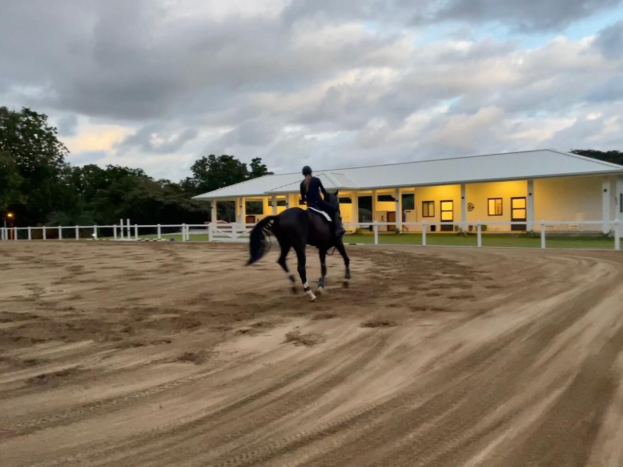 A person rides a horse in a dirt arena in front of a modern stable building — Las Lagartijas San Carlos, Cocle, FL