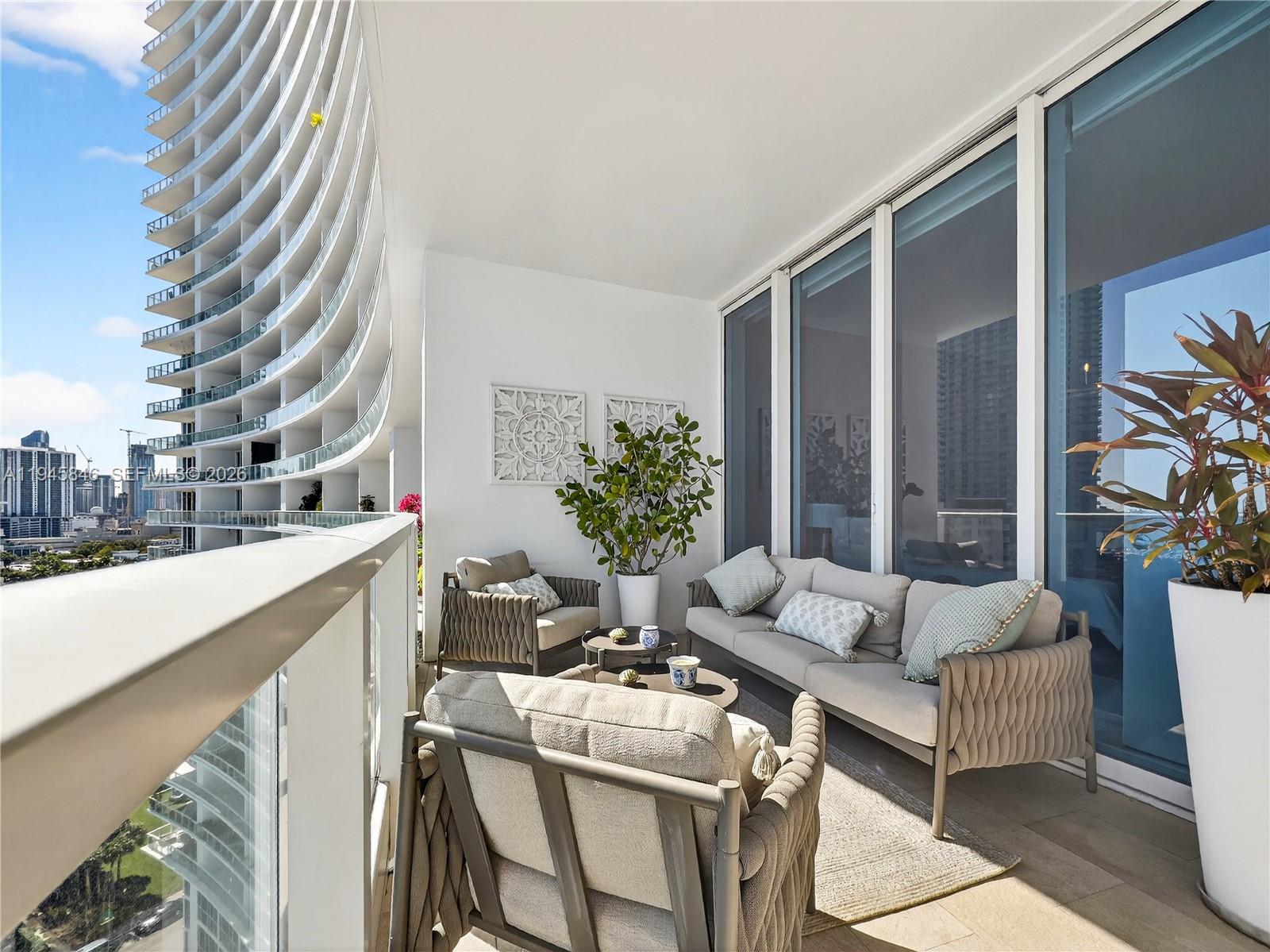 A furnished balcony features a curved glass railing, outdoor seating, and views of city skyscrapers — 2020 N Bayshore Dr # 1603, Miami FL 33137, MIAMI, FL
