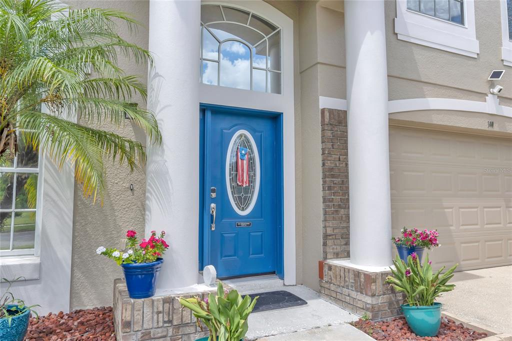 A blue front door with a decorative window is flanked by potted flowers and columns — 100 THORNBURY LANE, KISSIMMEE, FL