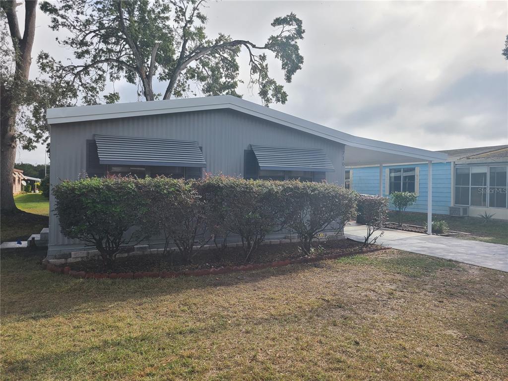 A gray mobile home features a carport, metal awnings, and a landscaped front yard — 710 COACHMAN DRIVE, LEESBURG, FL