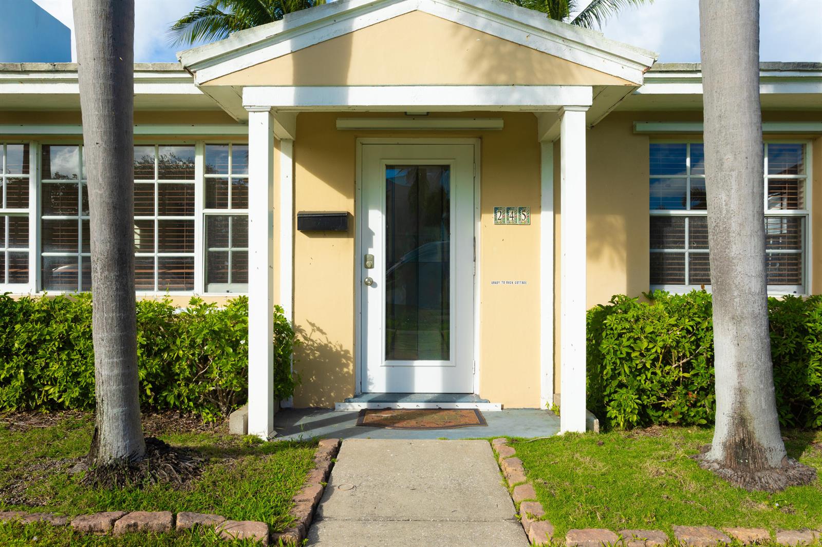 A beige cottage exterior features a white front door, a concrete walkway, and palm trees — 245 Cortez Road, West Palm Beach FL 33405, WEST PALM BEACH, FL