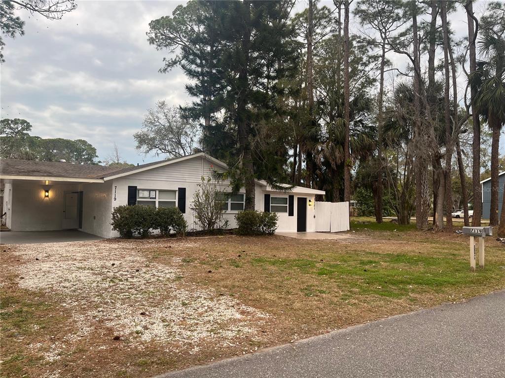 A white single-story house with a carport and a gravel driveway sits among tall trees — 7119-7121 OAKWOOD, NEW PORT RICHEY, FL