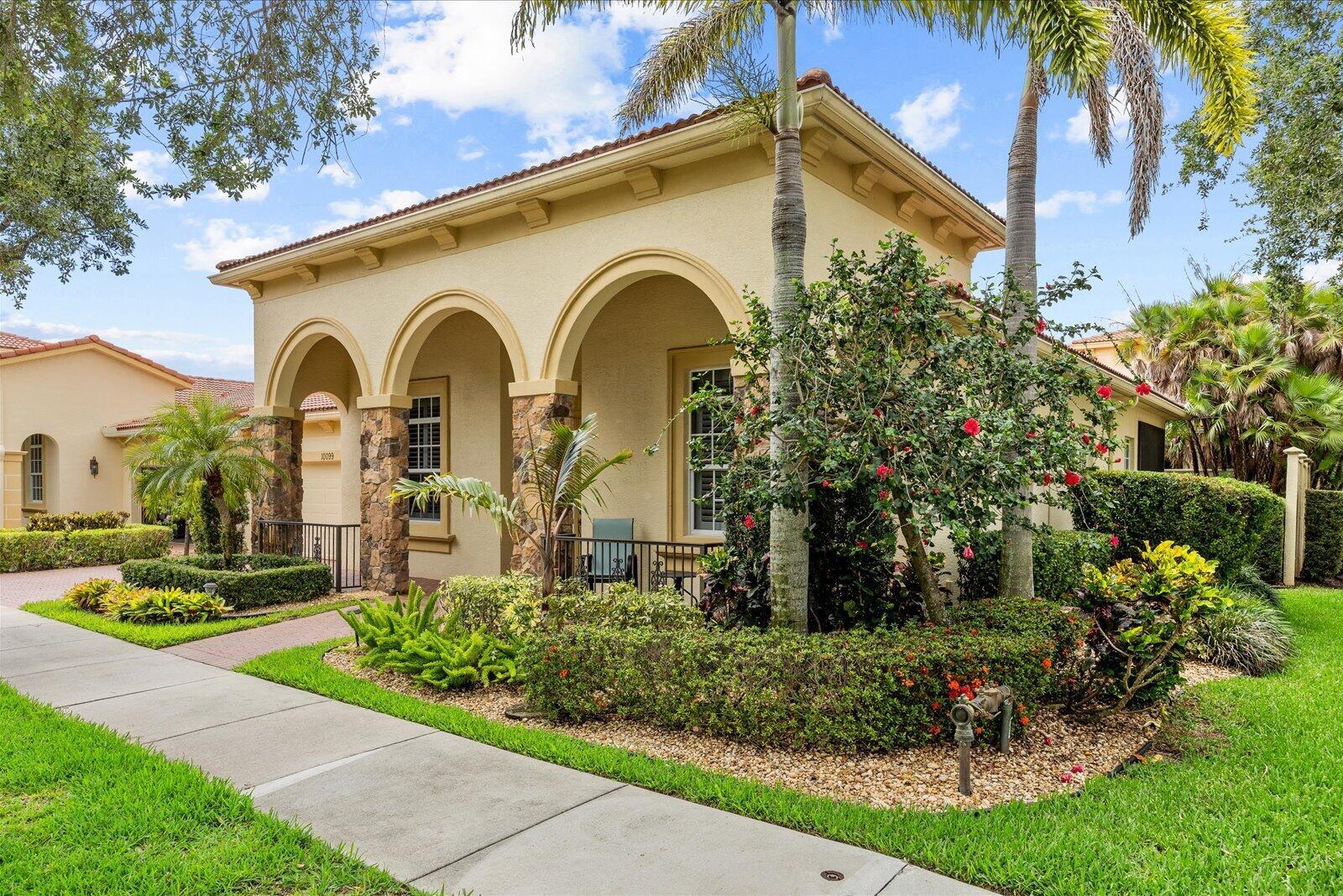 A beige stucco house with arched entryways, stone pillars, and lush landscaping under a blue sky — 10099 SW Duccio Lane, FL 34986, FL