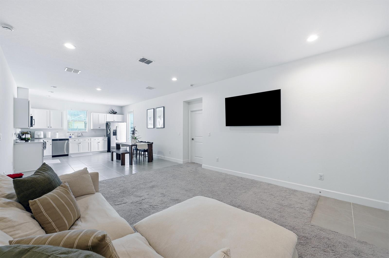 A living room with a wall-mounted television, gray carpet, and an adjacent open kitchen — 8452 Dorsey Street, Spring Hill FL 34608, SPRING HILL, FL