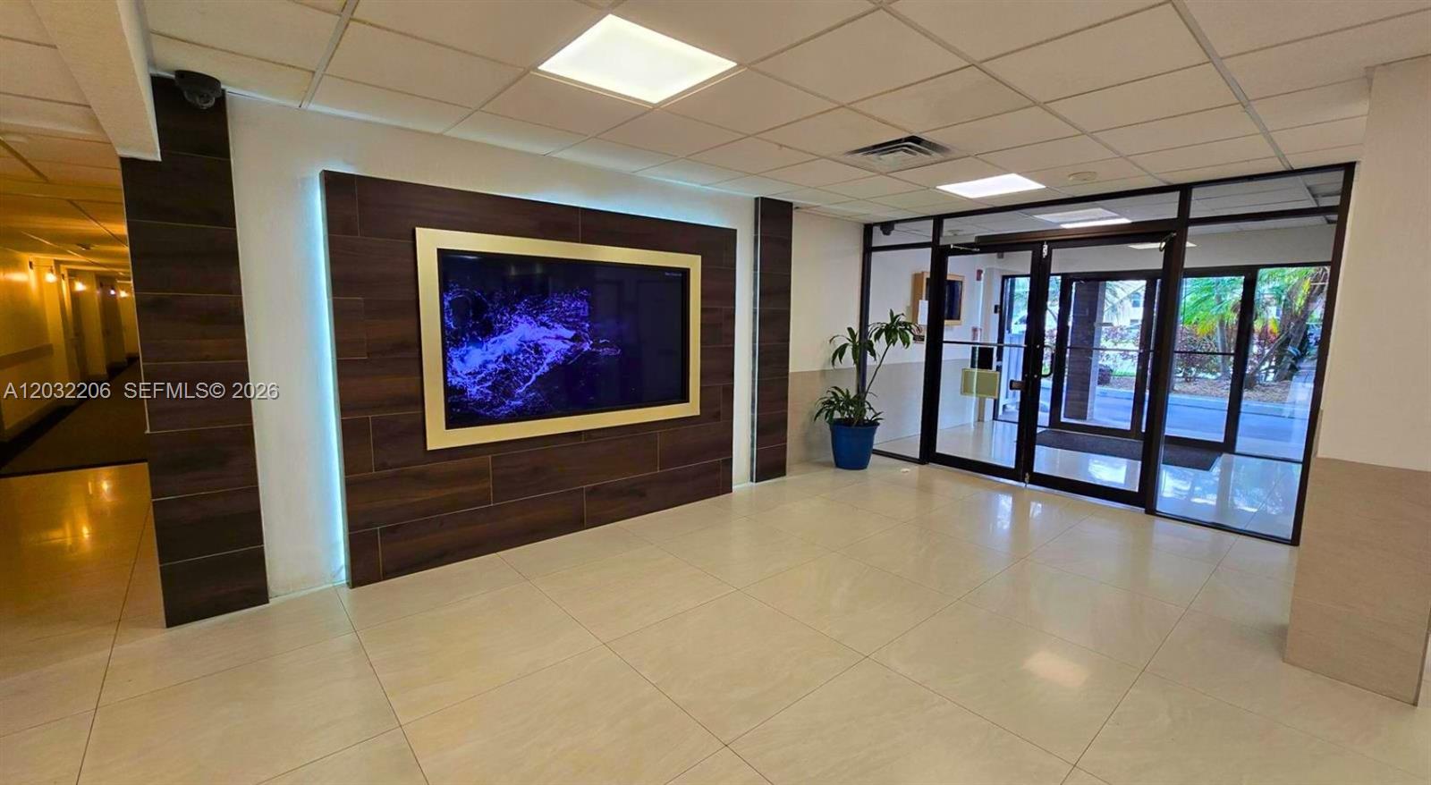 A modern lobby features a large wall-mounted television, tiled flooring, and glass-paneled entrance doors — 4164 Inverrary Dr # 204, Lauderhill FL 33319, LAUDERHILL, FL