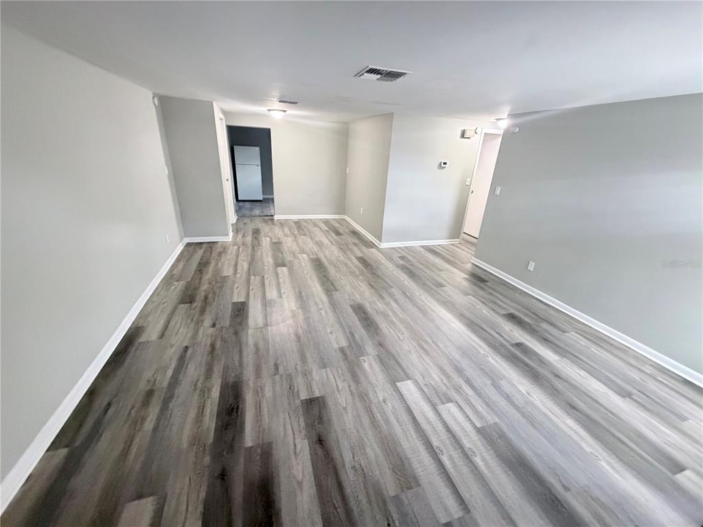 An empty room with gray walls and wood-look laminate flooring leads to a kitchen area — 8923 CANDLEWICK LANE, PORT RICHEY, FL