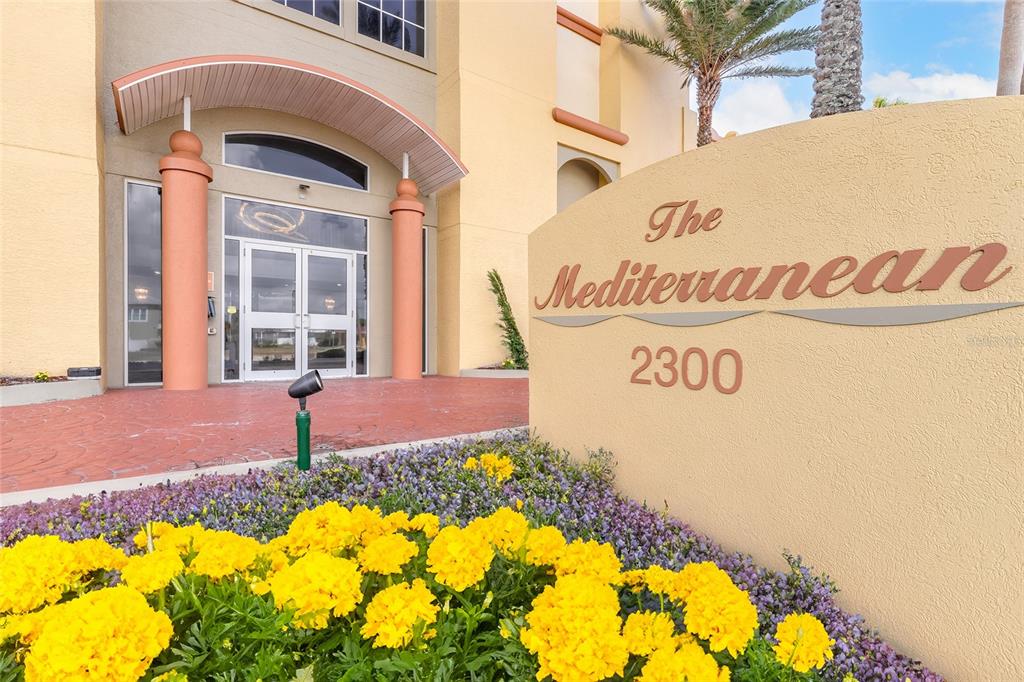 A sign for The Mediterranean 2300 stands in front of a building entrance with yellow flowers — 2300 N ATLANTIC AVENUE UNIT 301, DAYTONA BEACH, FL