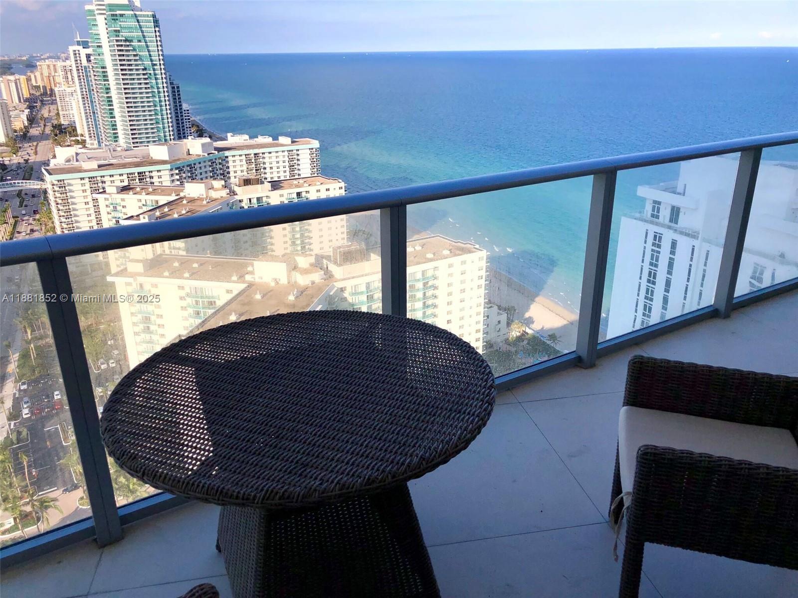 A balcony with a round wicker table and chair overlooks the ocean and city buildings — 4111 S Ocean Dr # 2910, Hollywood FL 33019, HOLLYWOOD, FL