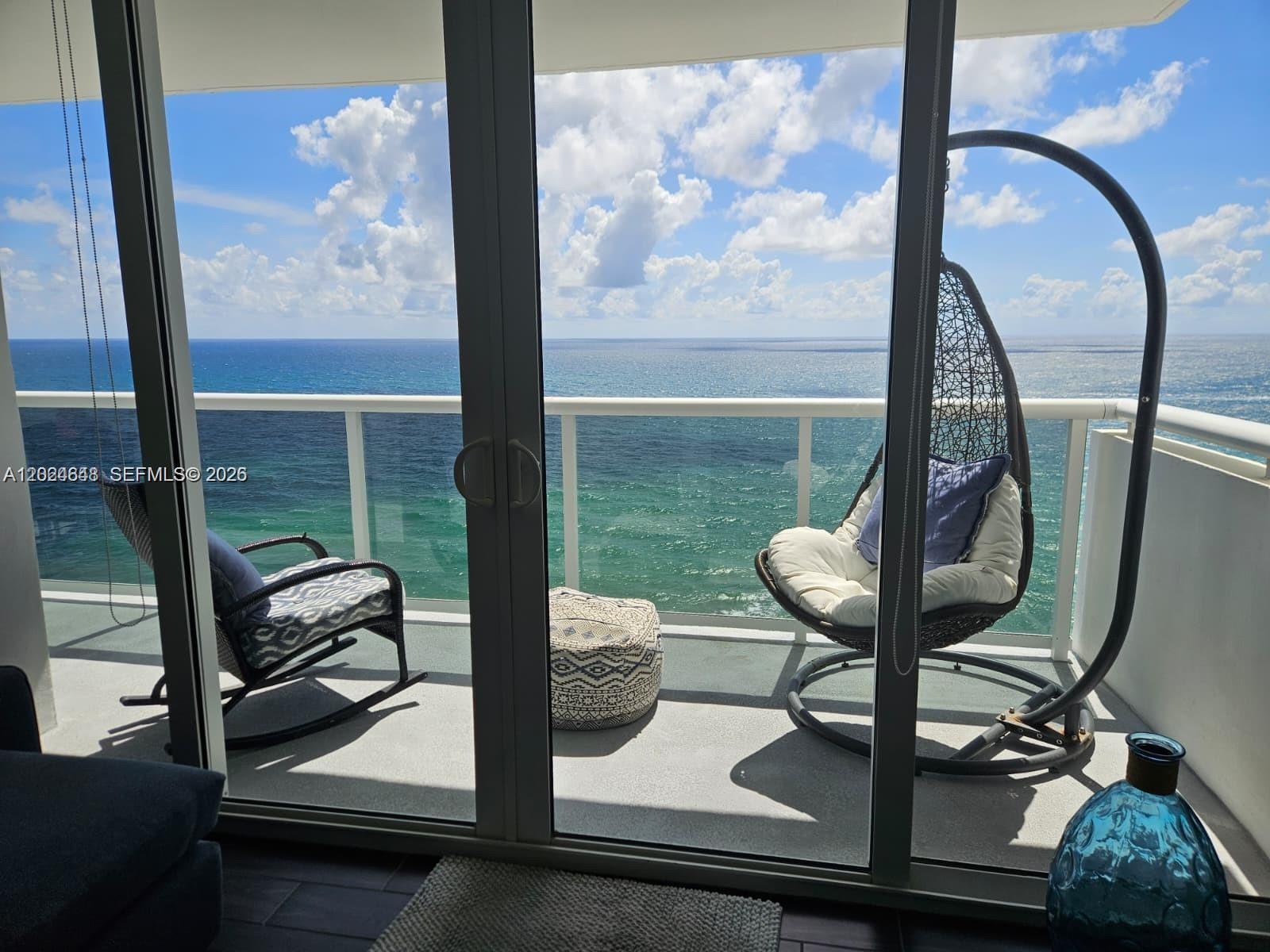 A balcony with a rocking chair and hanging chair overlooks the ocean through glass doors — 3140 S Ocean Dr # 1705, Hallandale Beach FL 33009, HALLANDALE BEACH, FL