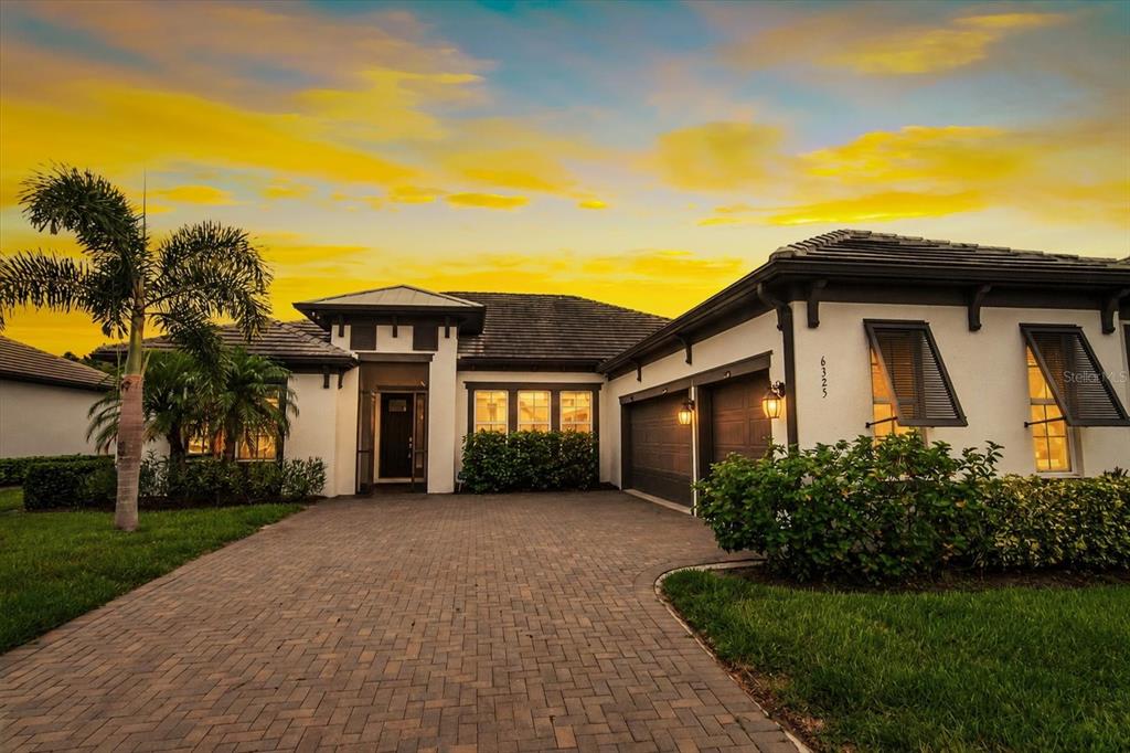 A single-story house with a paved driveway, dark garage doors, and shutters at sunset — 6325 RIVO LAKES BOULEVARD, SARASOTA, FL