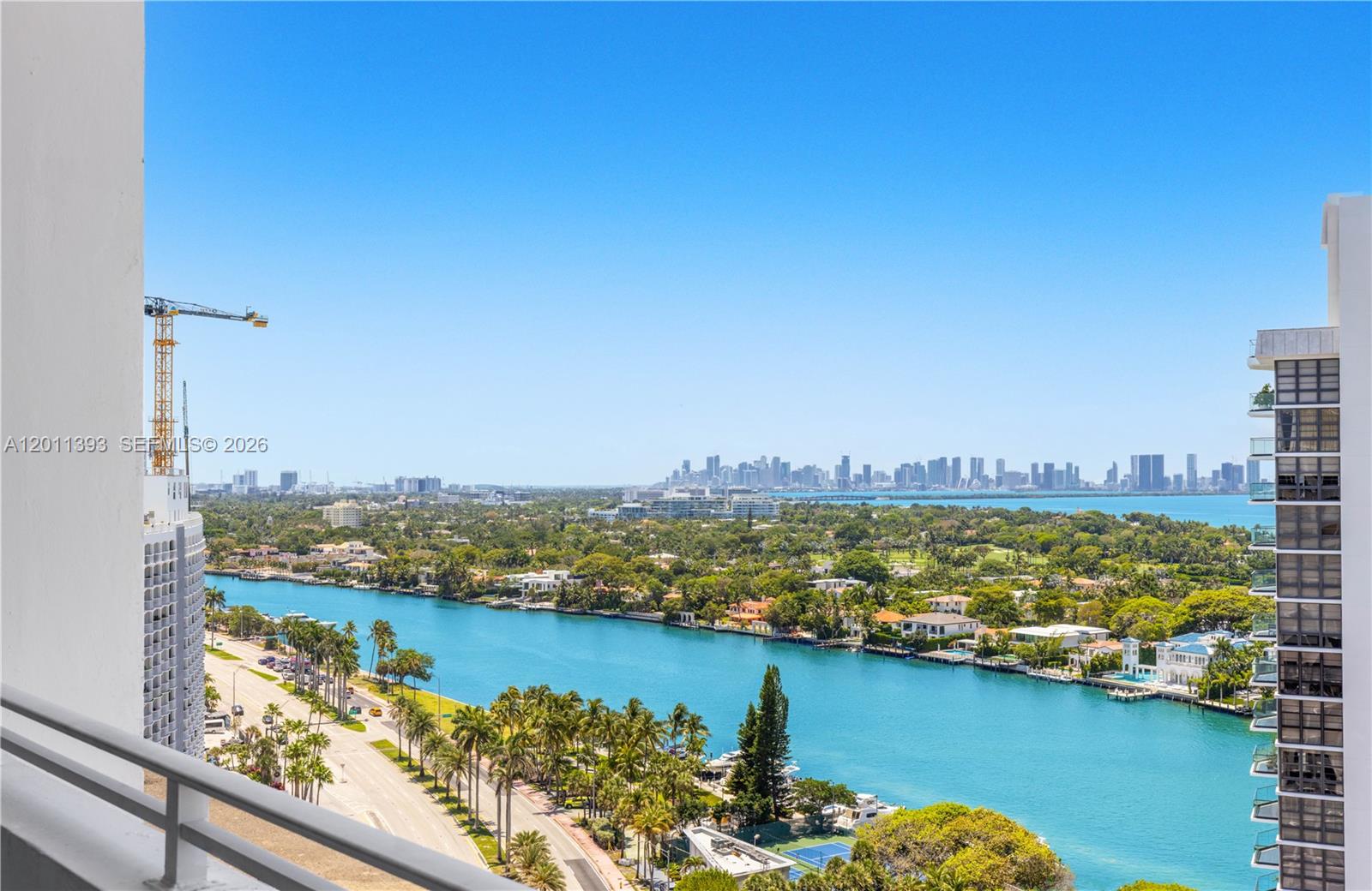 A high-angle view from a balcony overlooks a waterway, palm trees, and a distant skyline — 5445 Collins Ave, Miami Beach FL 33140, MIAMI BEACH, FL