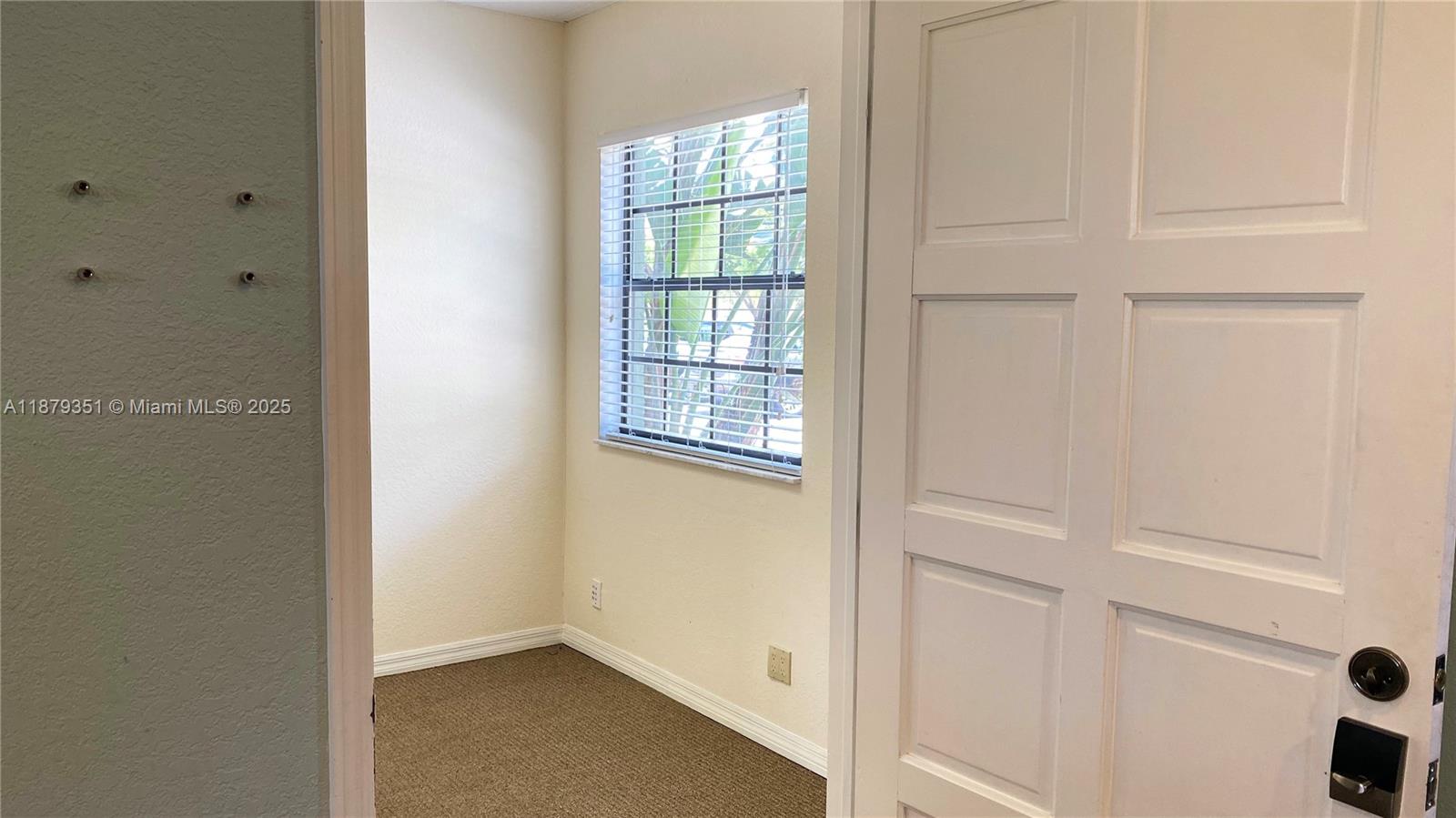A small room features beige walls, brown carpeting, a window with blinds, and a white door — 805 E Hillsboro Blvd # 102, Deerfield Beach FL 33441, DEERFIELD BEACH, FL