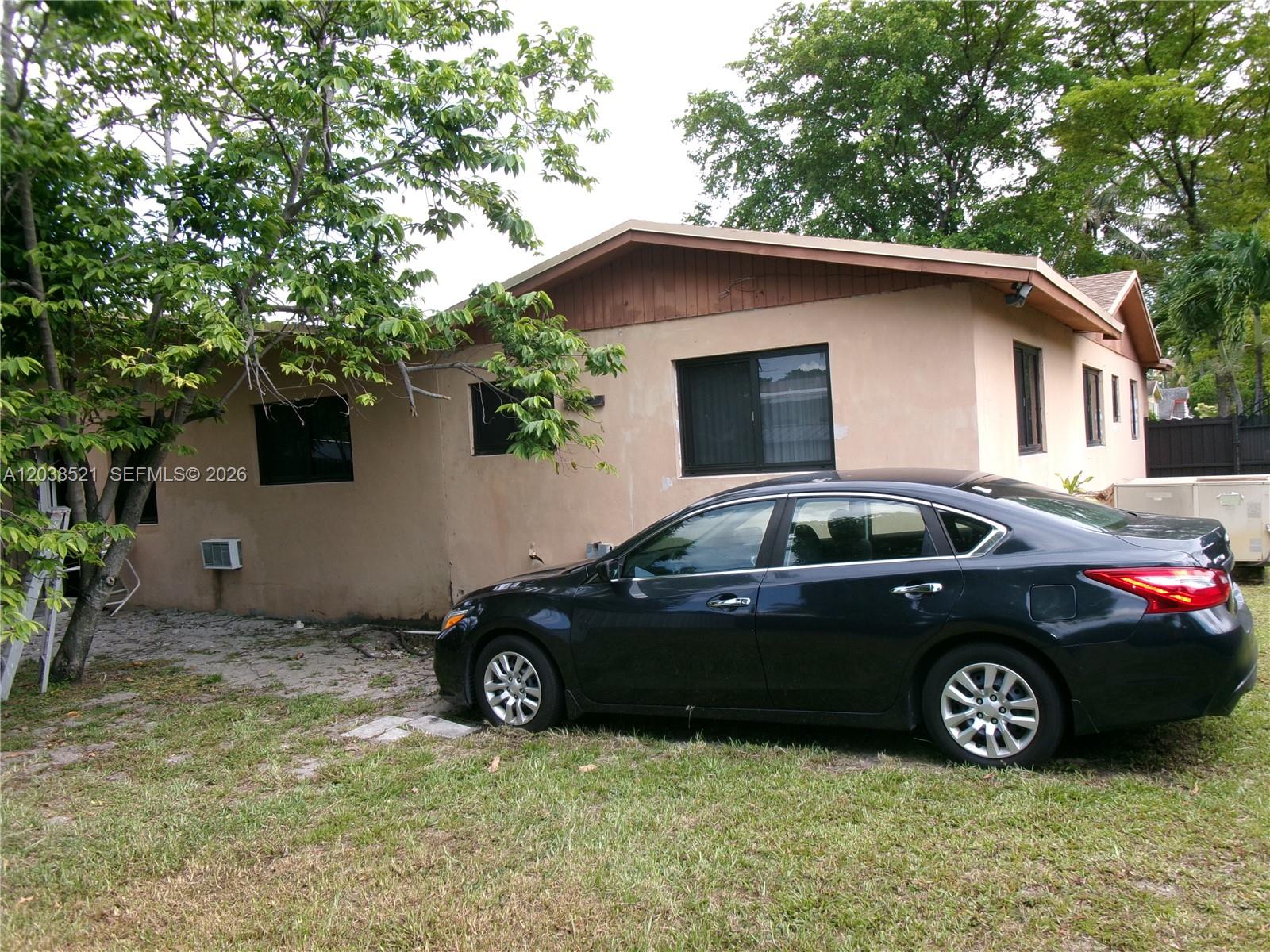 A dark sedan is parked on the grass in front of a tan, single-story house — MIAMI, FL
