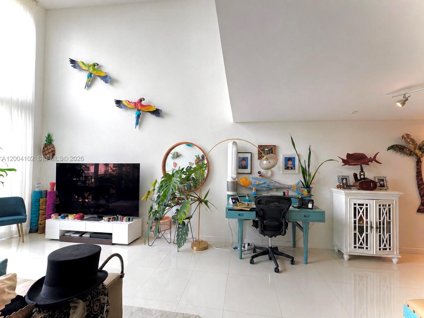 A living room features a television, a blue desk, wall-mounted parrot decor, and white tile — 6000 Collins Ave # 515, Miami Beach FL 33140, MIAMI BEACH, FL