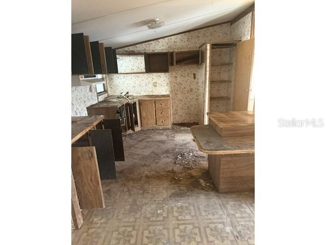 A dilapidated kitchen features wood cabinetry, patterned wallpaper, and damaged flooring — 36118 ZINNIA AVENUE, ZEPHYRHILLS, FL