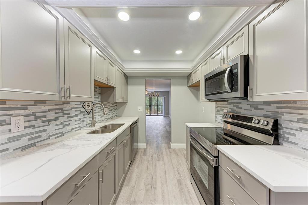 A galley kitchen features gray cabinets, white marble-look countertops, stainless steel appliances, and tile backsplash — 515 30TH AVENUE W UNIT H201, BRADENTON, FL