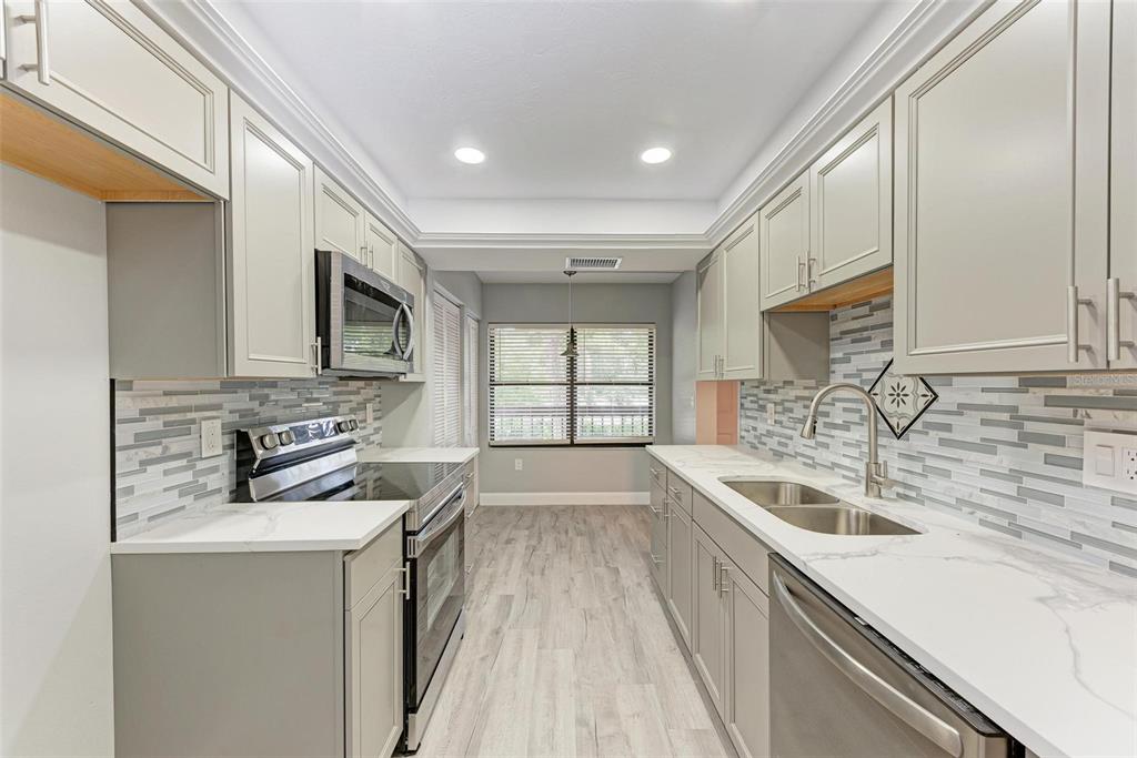 A galley kitchen features light gray cabinets, white countertops, a tiled backsplash, and stainless appliances — 515 30TH AVENUE W UNIT H201, BRADENTON, FL