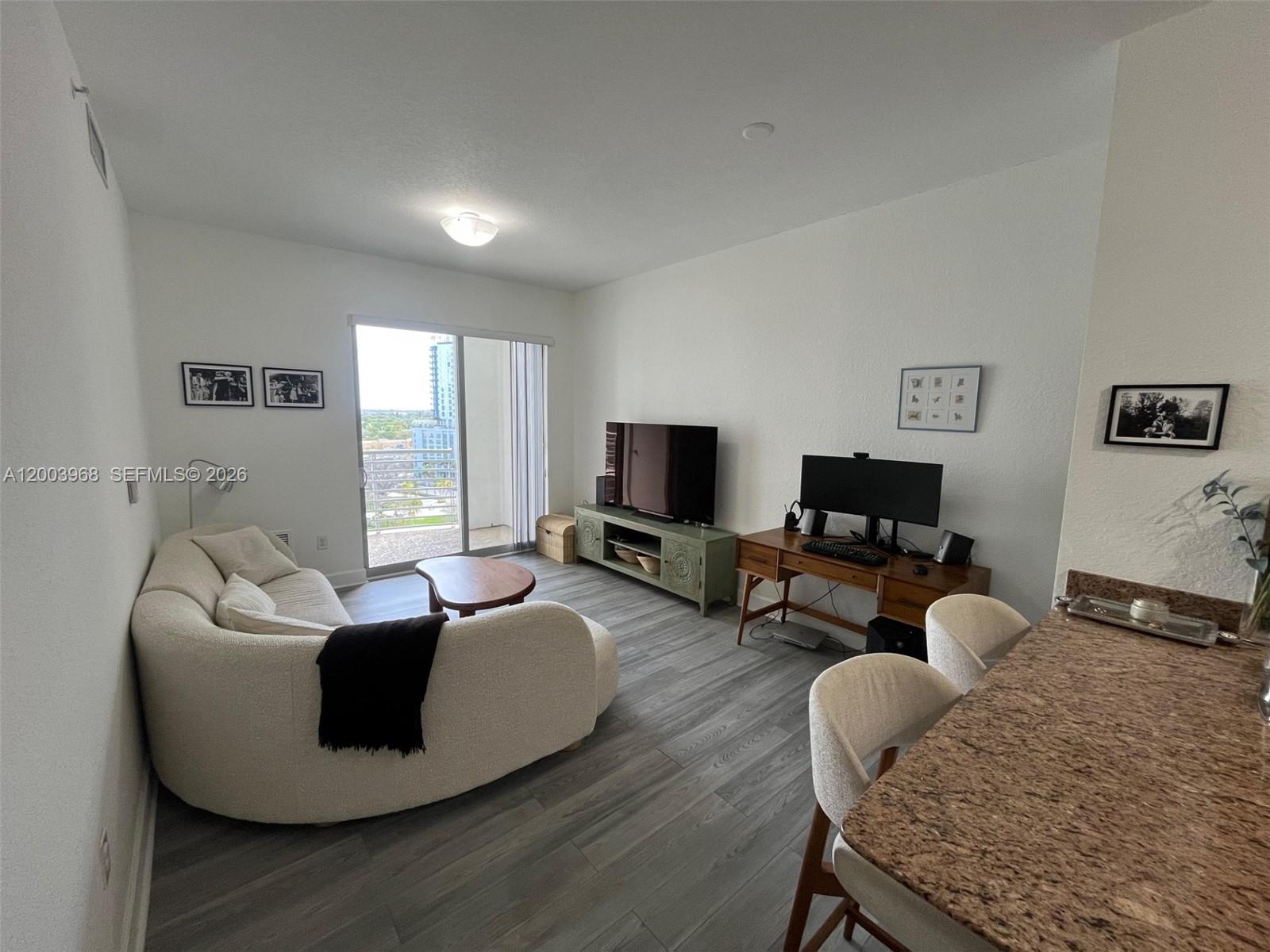 A living room features a curved sofa, television console, desk, and sliding glass balcony doors — 1830 Radius Dr # 1218, Hollywood FL 33020, HOLLYWOOD, FL