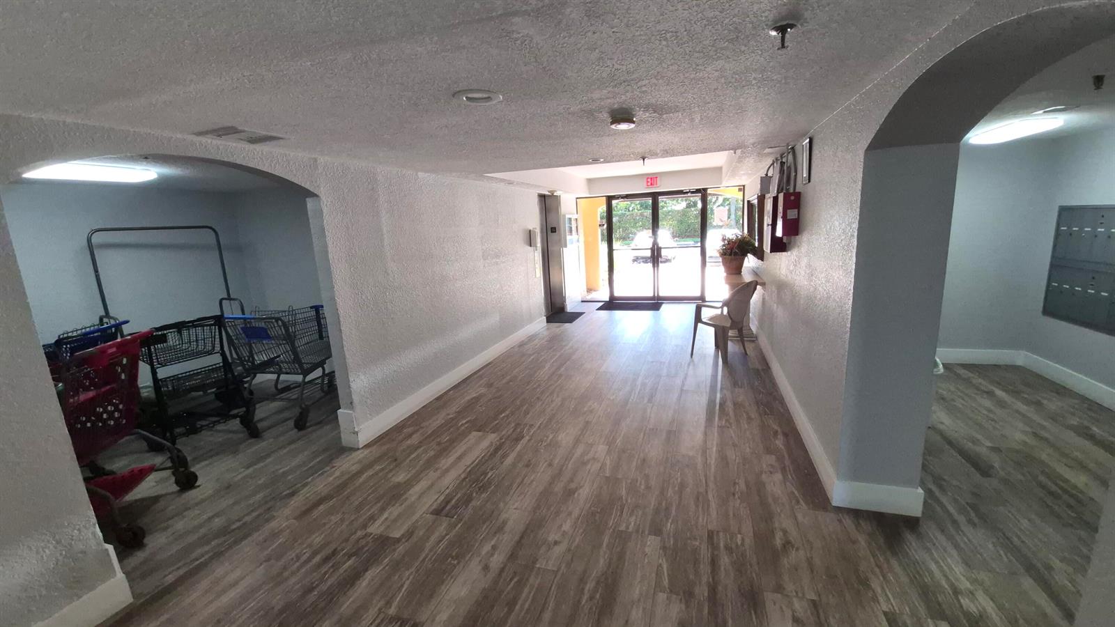 A hallway with wood-look flooring leads to a glass exit door with shopping carts nearby — 450 Egret Circle # 9403, Delray Beach FL 33444, DELRAY BEACH, FL