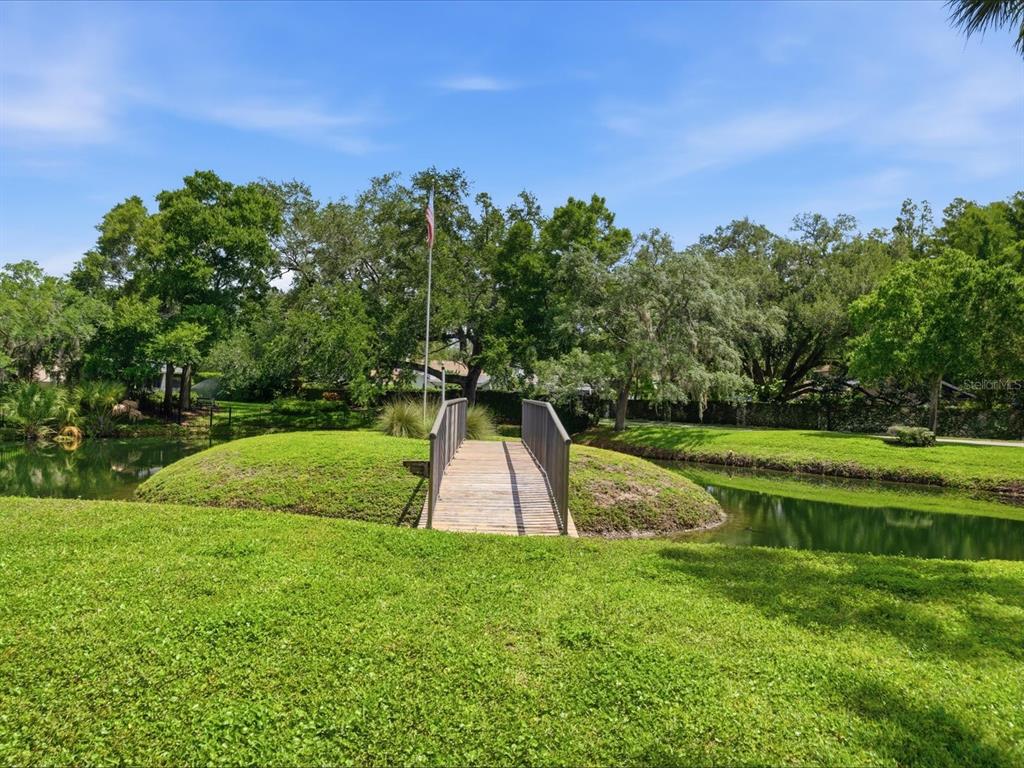 A wooden footbridge crosses a small pond in a grassy, tree-lined outdoor area — 13626 GREENFIELD DRIVE UNIT 401, TAMPA, FL