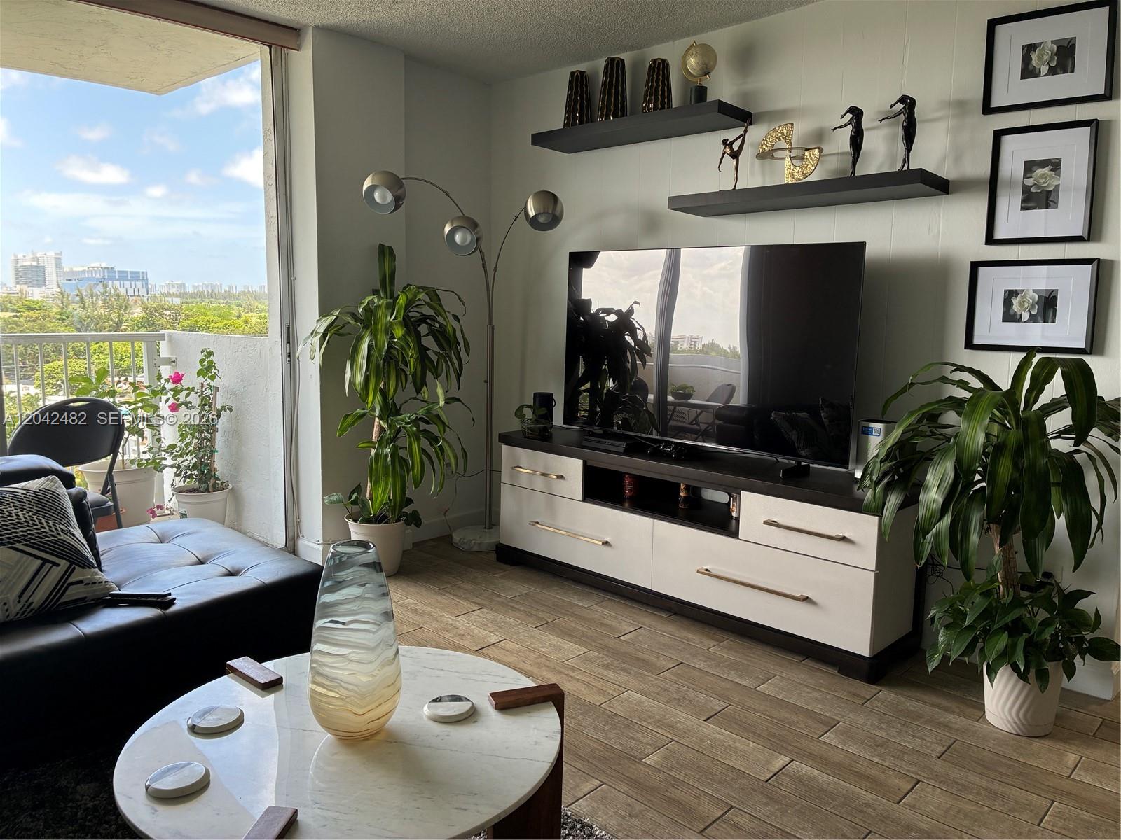 A living room features a television on a console, floating shelves, plants, and a balcony — 2000 NE 135th St # 710, North Miami FL 33181, NORTH MIAMI, FL
