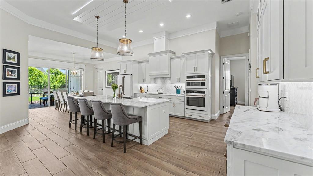 A modern white kitchen features a marble-topped island, gray bar stools, and wood-look tile flooring — 6509 HERITAGE PARK PLACE, LAKELAND, FL