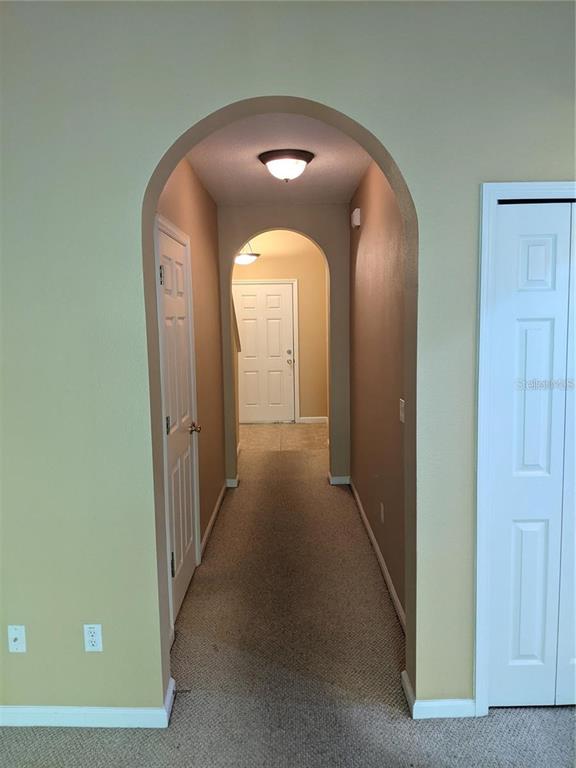 A hallway with arched entryways, beige walls, and carpeted flooring leads to a white door — 10272 PARK COMMONS DRIVE, ORLANDO, FL