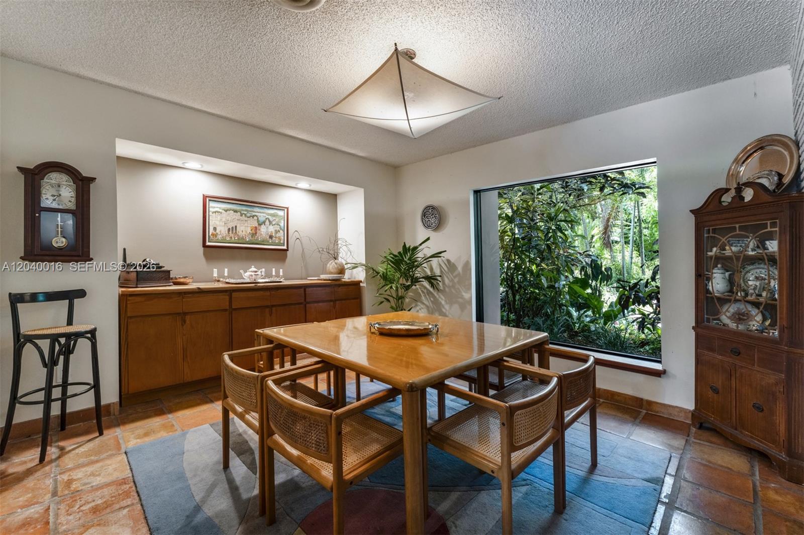 A dining room features a square wooden table, cane chairs, tile floors, and a large window — 11100 SW 67th Ave, Pinecrest FL 33156, PINECREST, FL