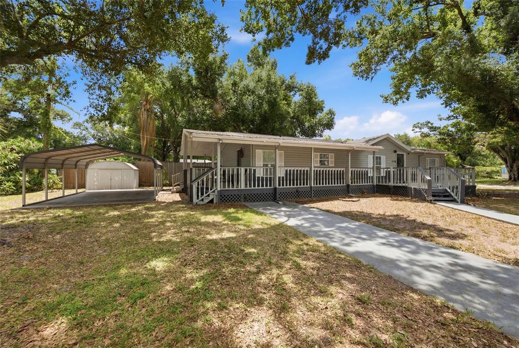 A single-story manufactured home with a front porch, carport, and shed under large trees — 5203 N MENDENHALL DRIVE, TAMPA, FL