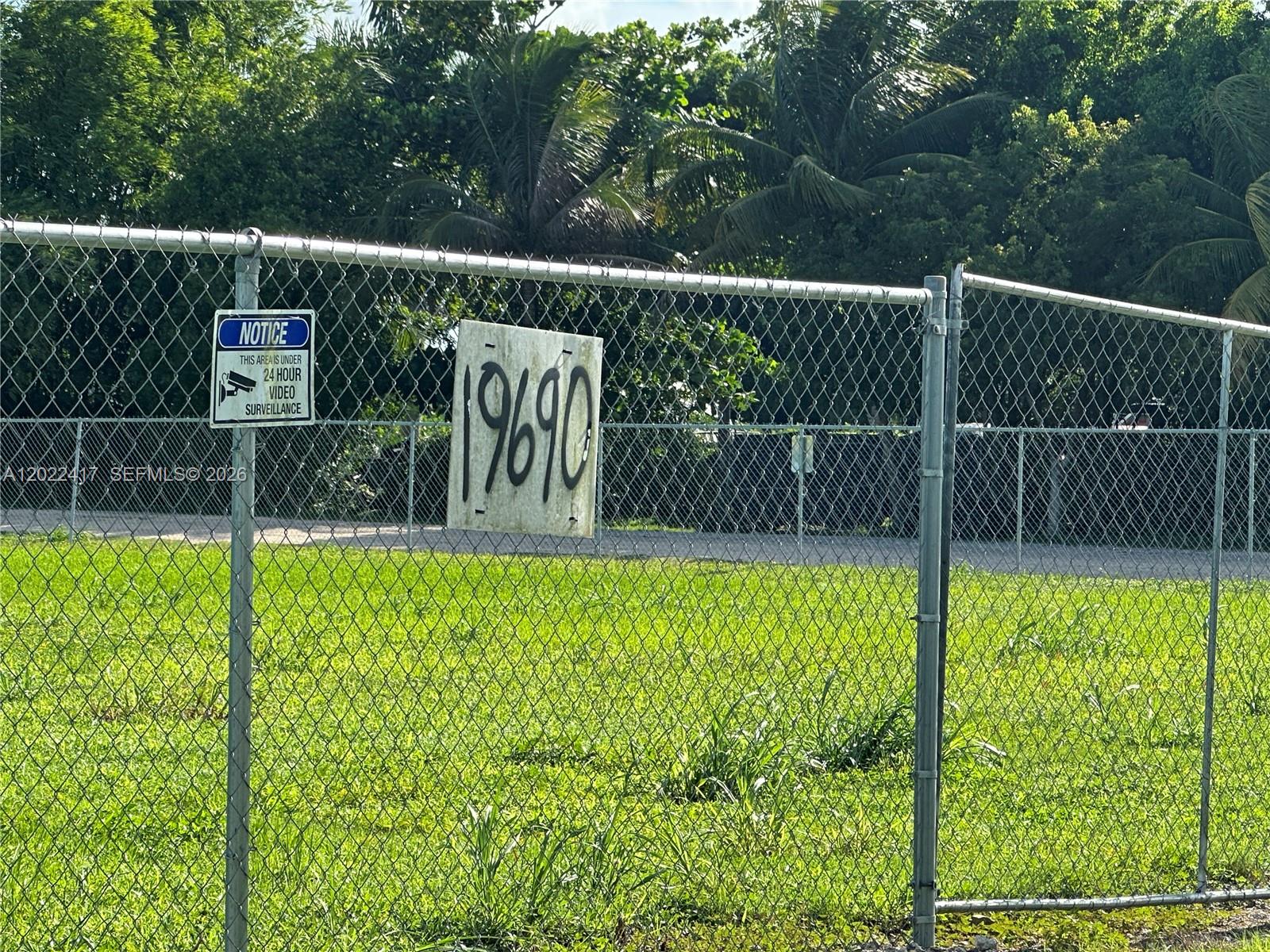 A chain-link fence surrounds a grassy lot with a surveillance sign and a house number — 19690 SW 206th St, Miami FL 33187, MIAMI, FL