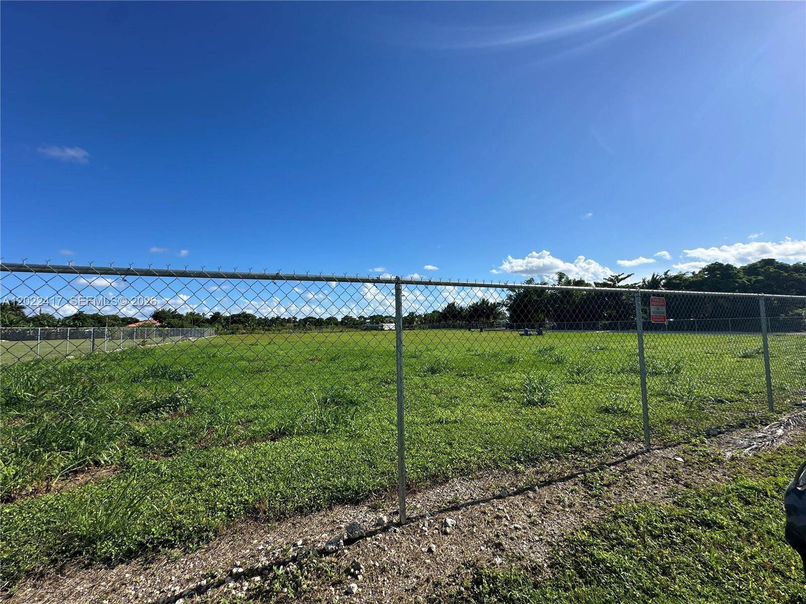 A chain-link fence encloses a large, grassy, undeveloped plot of land under a clear sky — 19690 SW 206th St, Miami FL 33187, MIAMI, FL
