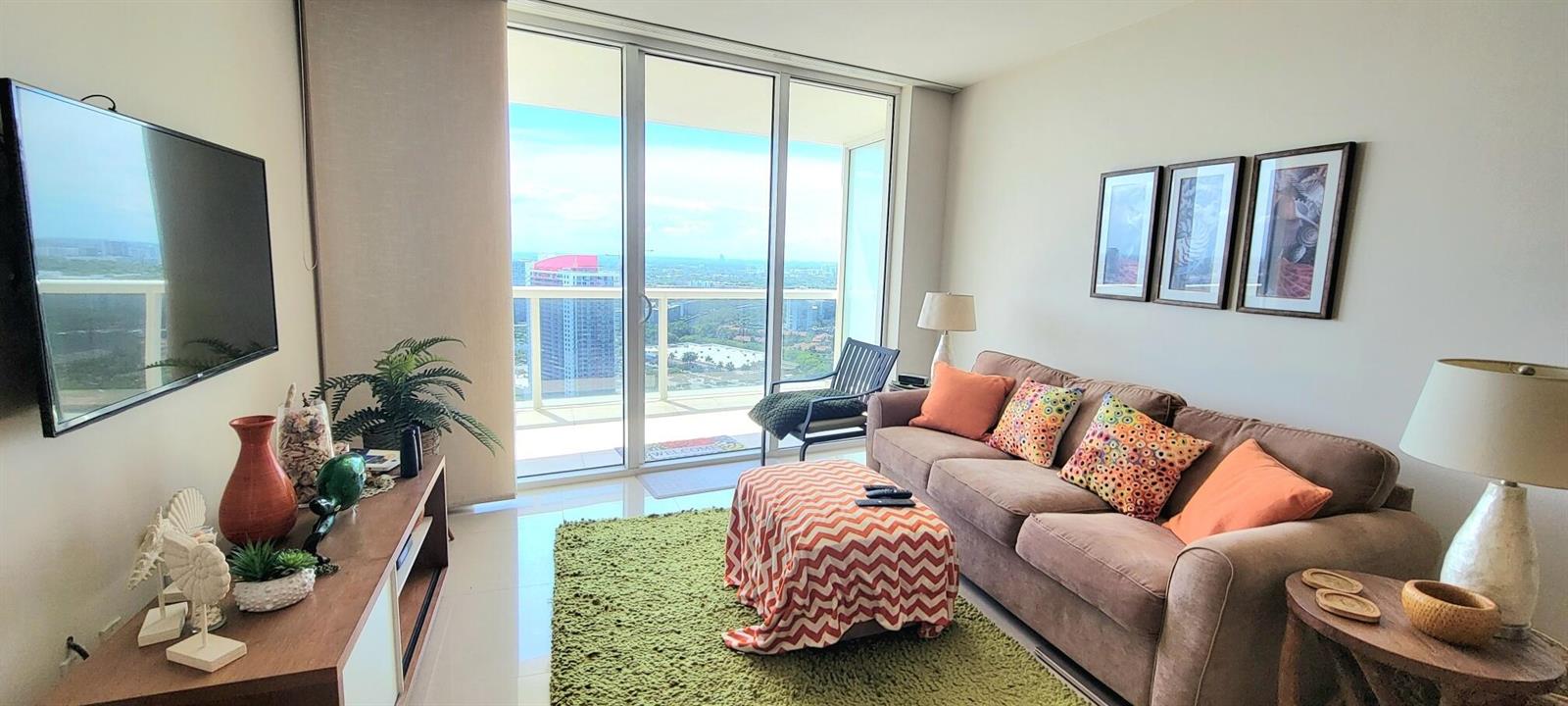 A living room features a brown sofa, green rug, television, and a balcony with city views — 1850 S Ocean Drive # 3806, Hallandale Beach FL 33009, HALLANDALE BEACH, FL