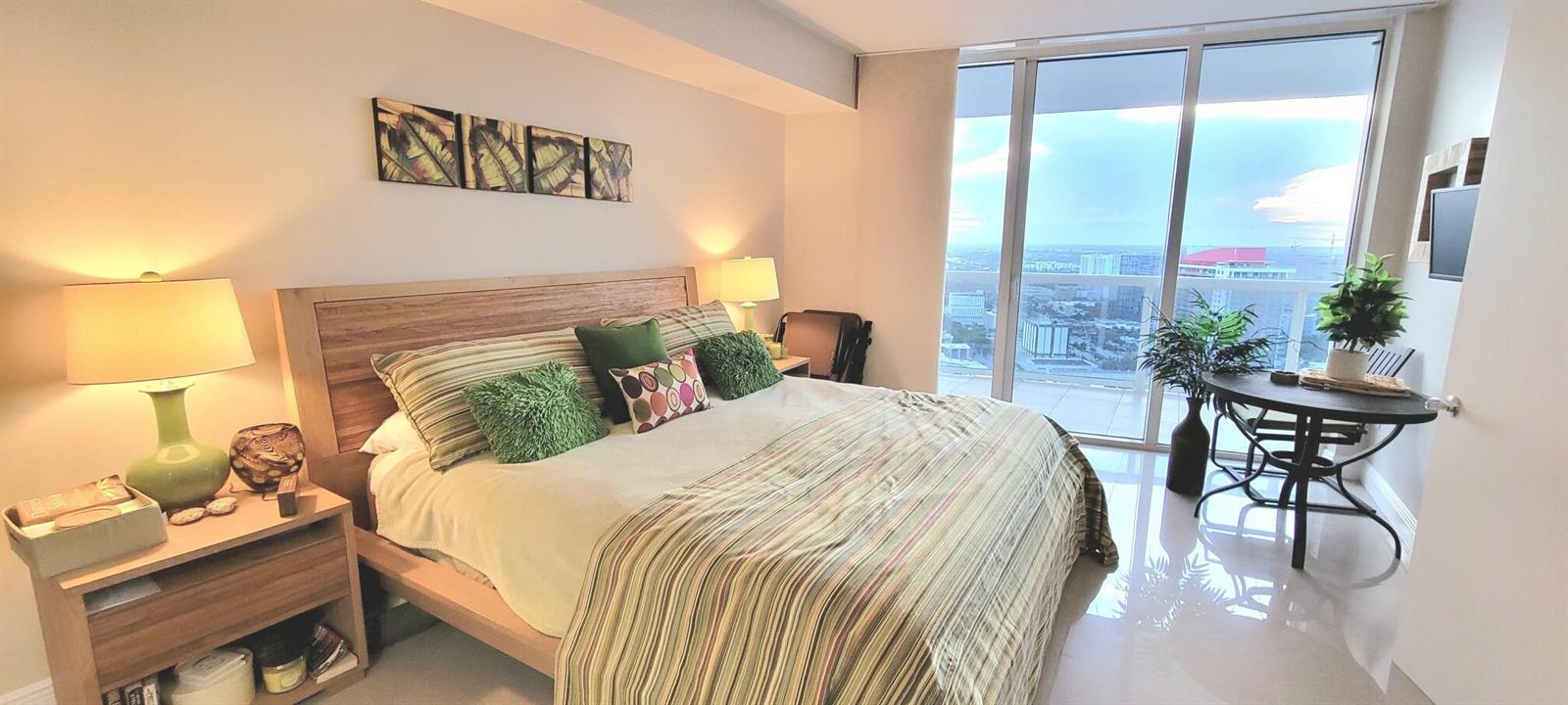 A bedroom features a wooden bed frame, green accents, and floor-to-ceiling windows with city views — 1850 S Ocean Drive # 3806, Hallandale Beach FL 33009, HALLANDALE BEACH, FL