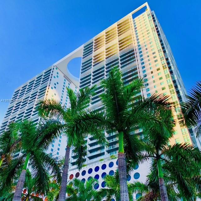 A low-angle view of a modern high-rise building with a circular cutout and palm trees — 500 Brickell Ave # 2208, Miami FL 33131, MIAMI, FL