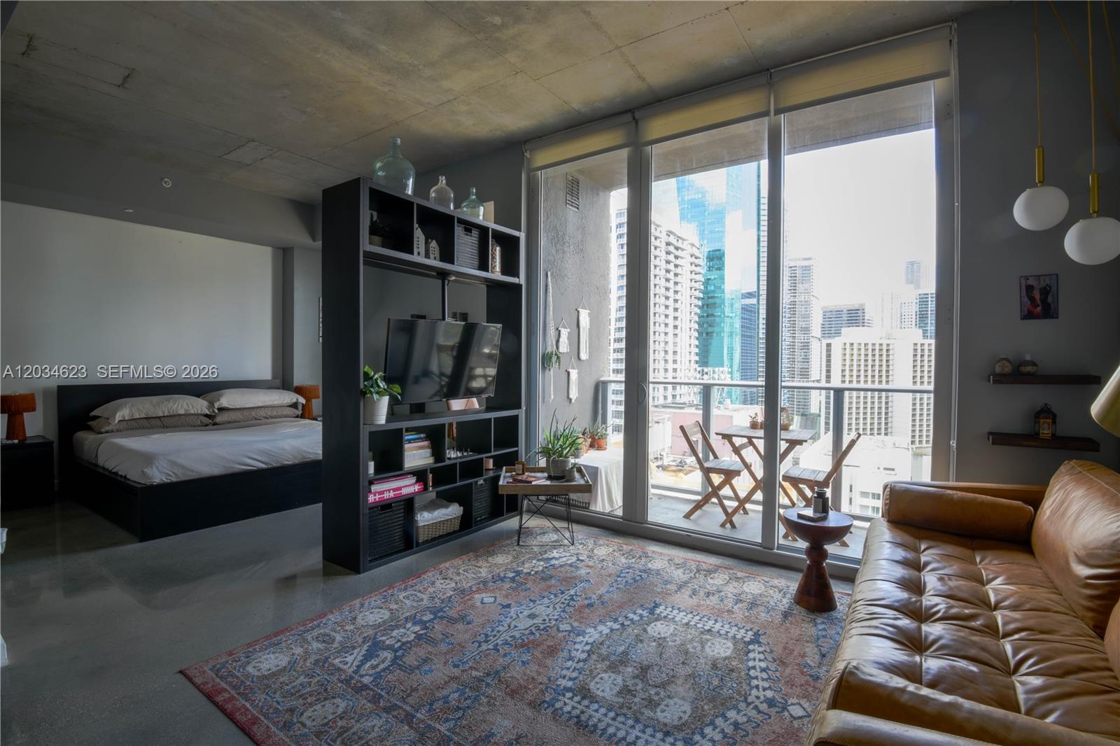A studio apartment features a concrete ceiling, a room-dividing bookshelf, and a balcony with city views — 151 SE 1st St # 1907, Miami FL 33131, MIAMI, FL