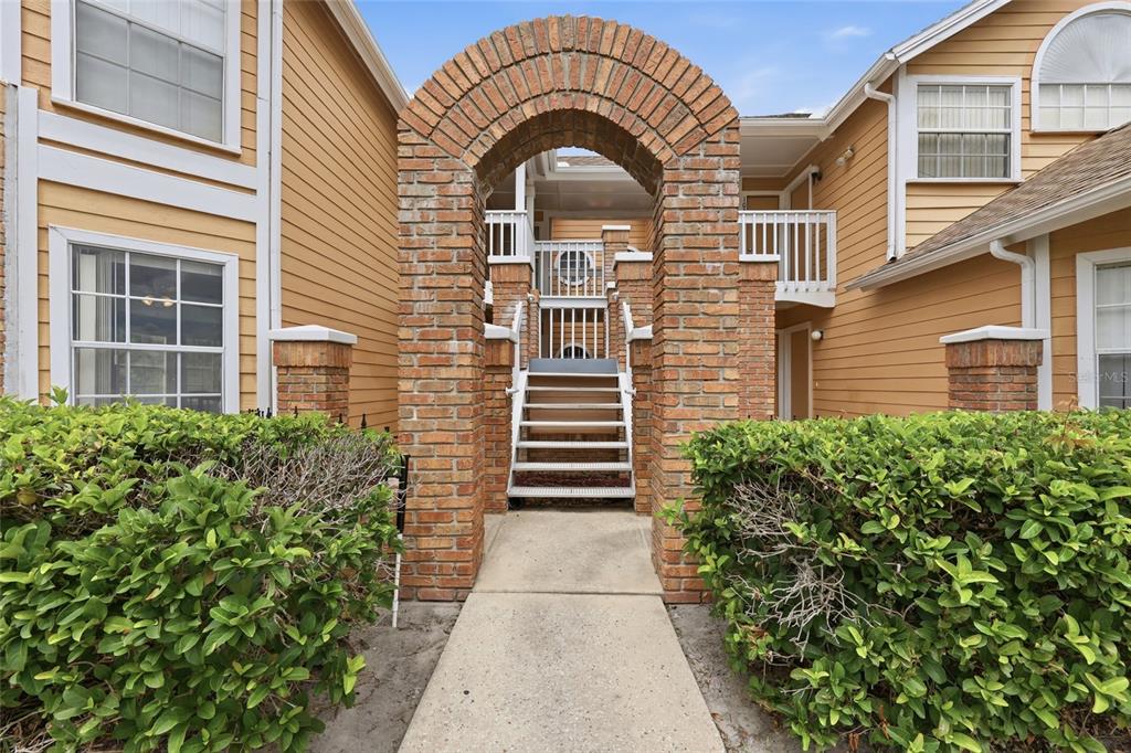 A brick archway leads to a staircase between two tan-sided residential buildings with green hedges — 2474 SWEETWATER CLUB CIRCLE UNIT 101, KISSIMMEE, FL