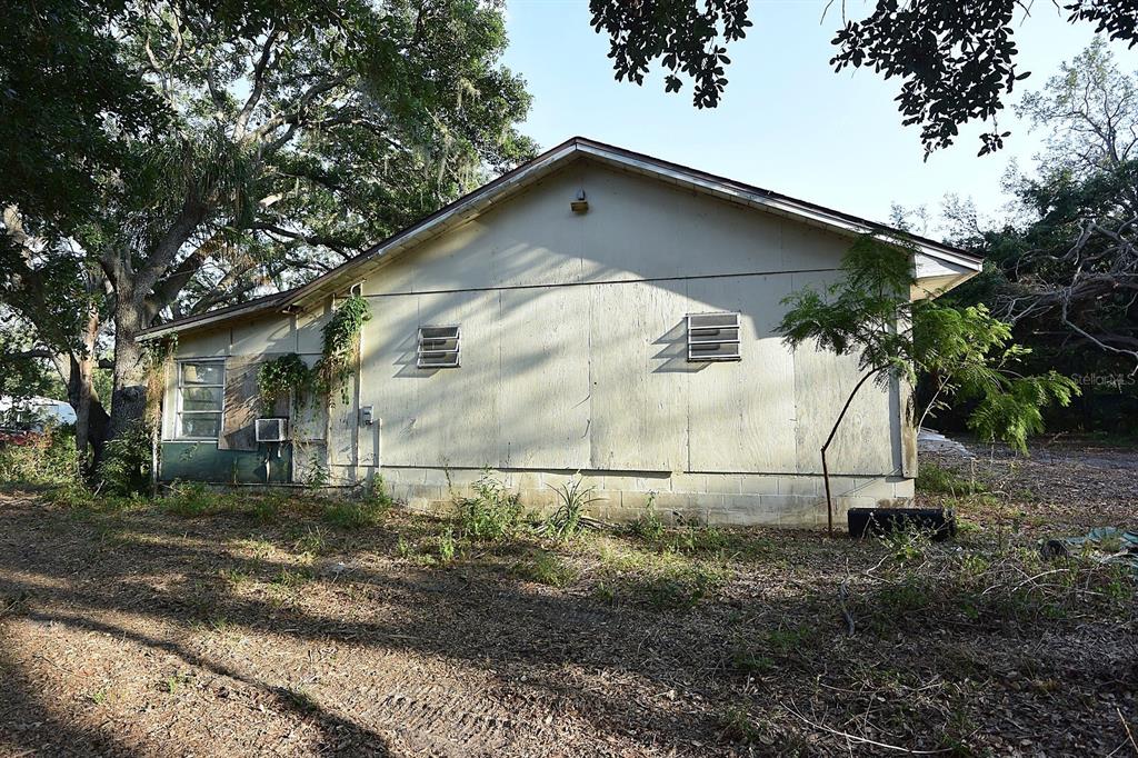 A side view of a single-story house with plywood siding, louvered vents, and overgrown landscaping — 7610 ANNA AVENUE, GIBSONTON, FL