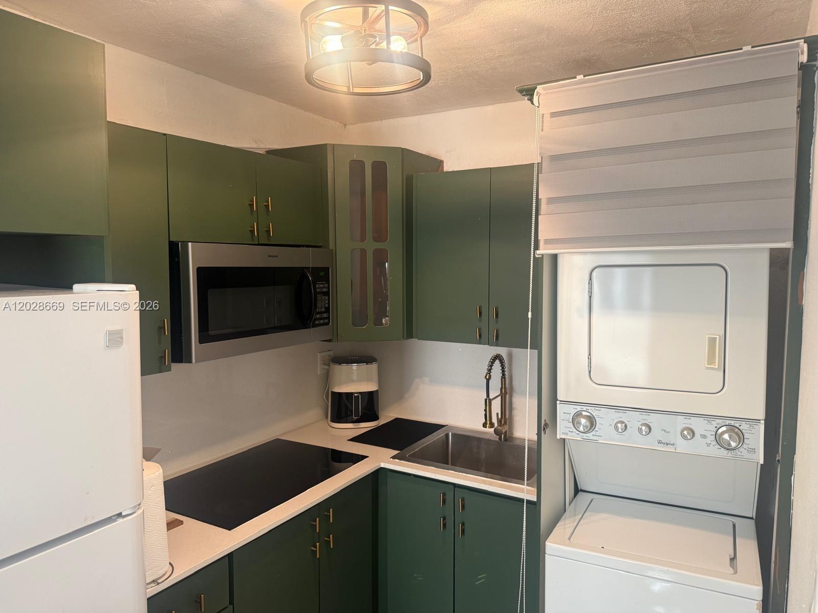 A small kitchen features green cabinets, stainless steel appliances, a sink, and a stacked washer-dryer — 7928 Harding Ave # 2, Miami Beach FL 33141, MIAMI BEACH, FL