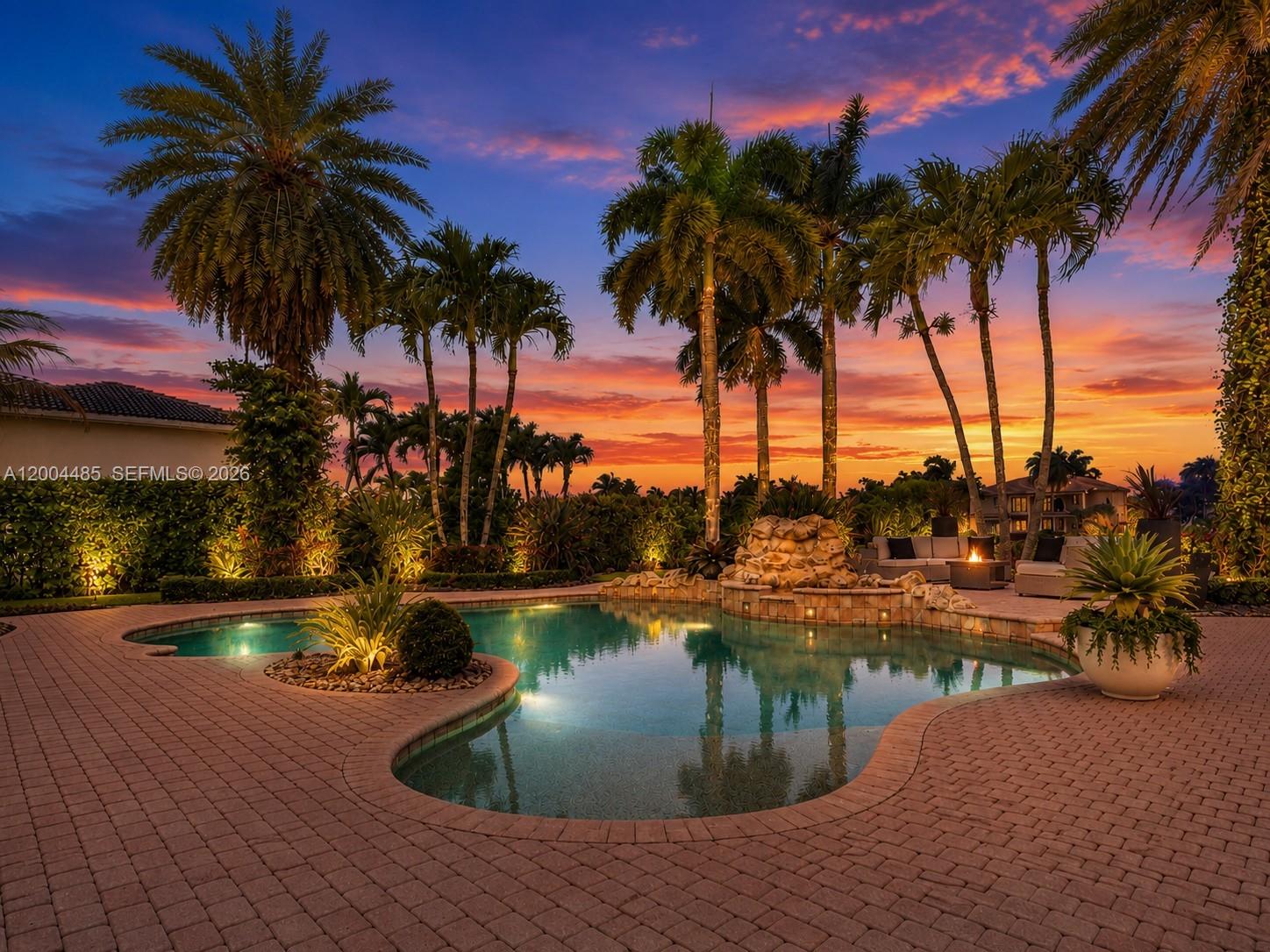 A swimming pool with a rock waterfall feature is surrounded by palm trees at sunset — 14857 SW 38th St, Davie FL 33331, DAVIE, FL