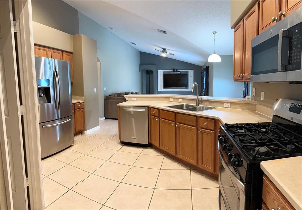 A kitchen with wood cabinets, stainless steel appliances, and tile flooring overlooks an open living area — 16016 BLOSSOM HILL LOOP, CLERMONT, FL