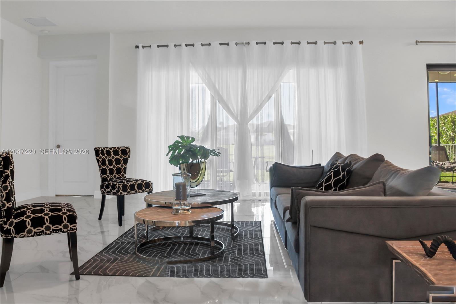 A living room features a grey sofa, patterned chairs, nesting coffee tables, and sheer curtains — 15750 Longboat Key Dr, Westlake FL 33470, WESTLAKE, FL