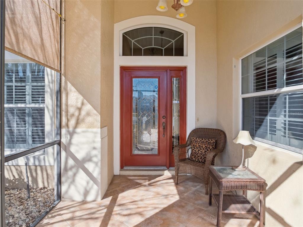 A covered entryway features a red front door, tiled floor, wicker chair, and side table — 20639 PRINCE RANIER PLACE, LEESBURG, FL