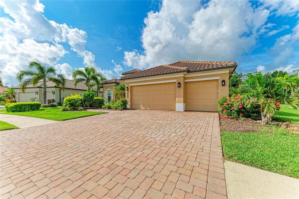 A tan house with a two-car garage and a brick paver driveway under a blue sky — 13440 CAMPANILE COURT, VENICE, FL