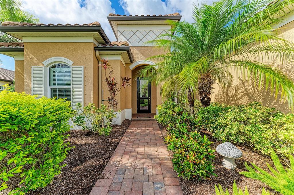A brick walkway leads to the front entrance of a tan house with palm trees — 13440 CAMPANILE COURT, VENICE, FL