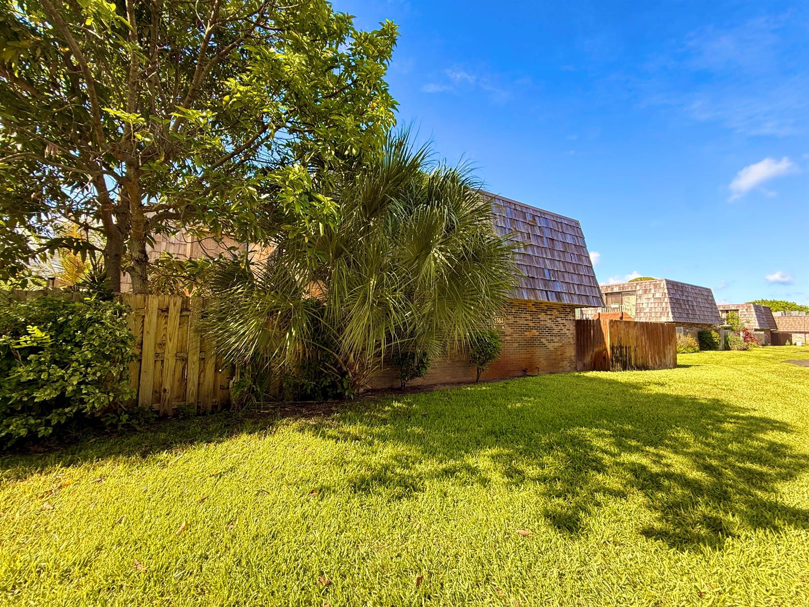 A brick building with a shingled roof stands behind a wooden fence and palm tree — 1684 Forest Lakes Circle # A, West Palm Beach FL 33406, WEST PALM BEACH, FL