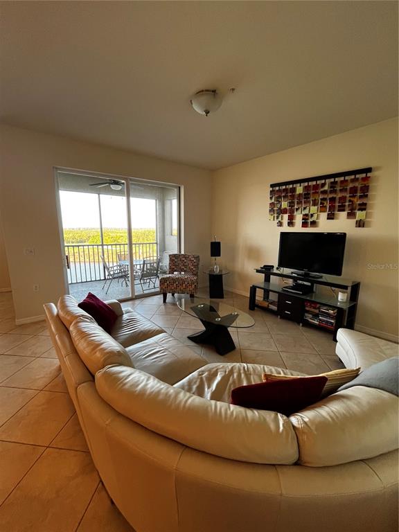 A living room features a large curved sofa, a television on a stand, and a balcony — 7911 GRAND ESTUARY TRAIL UNIT 406, BRADENTON, FL