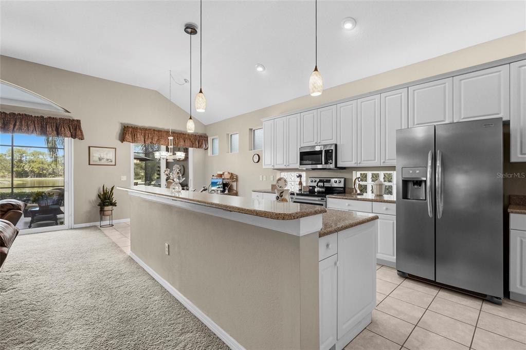 A kitchen with white cabinets, granite countertops, and stainless steel appliances overlooks a carpeted living area — 7822 TROON COURT, LAKEWOOD RANCH, FL